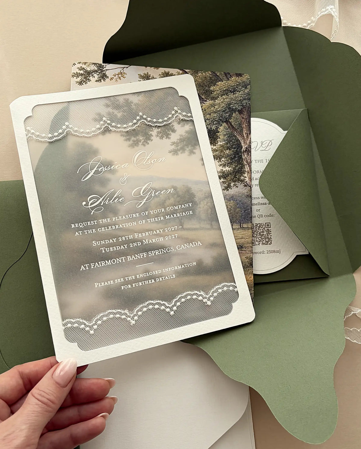 Hand lifting the lace-trimmed vellum wedding invitation out of the olive green four-petal folder, with the landscape print behind