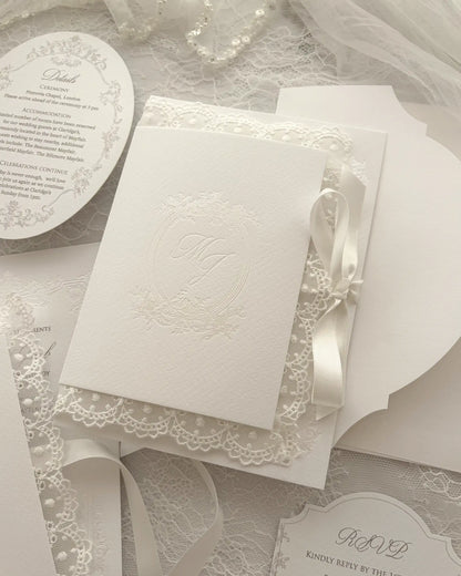 Alyson Lace White wedding invitation suite with embossed monogram card, lace trim, ribbon, RSVP card and envelope on delicate lace fabric background — Polina Perri. 