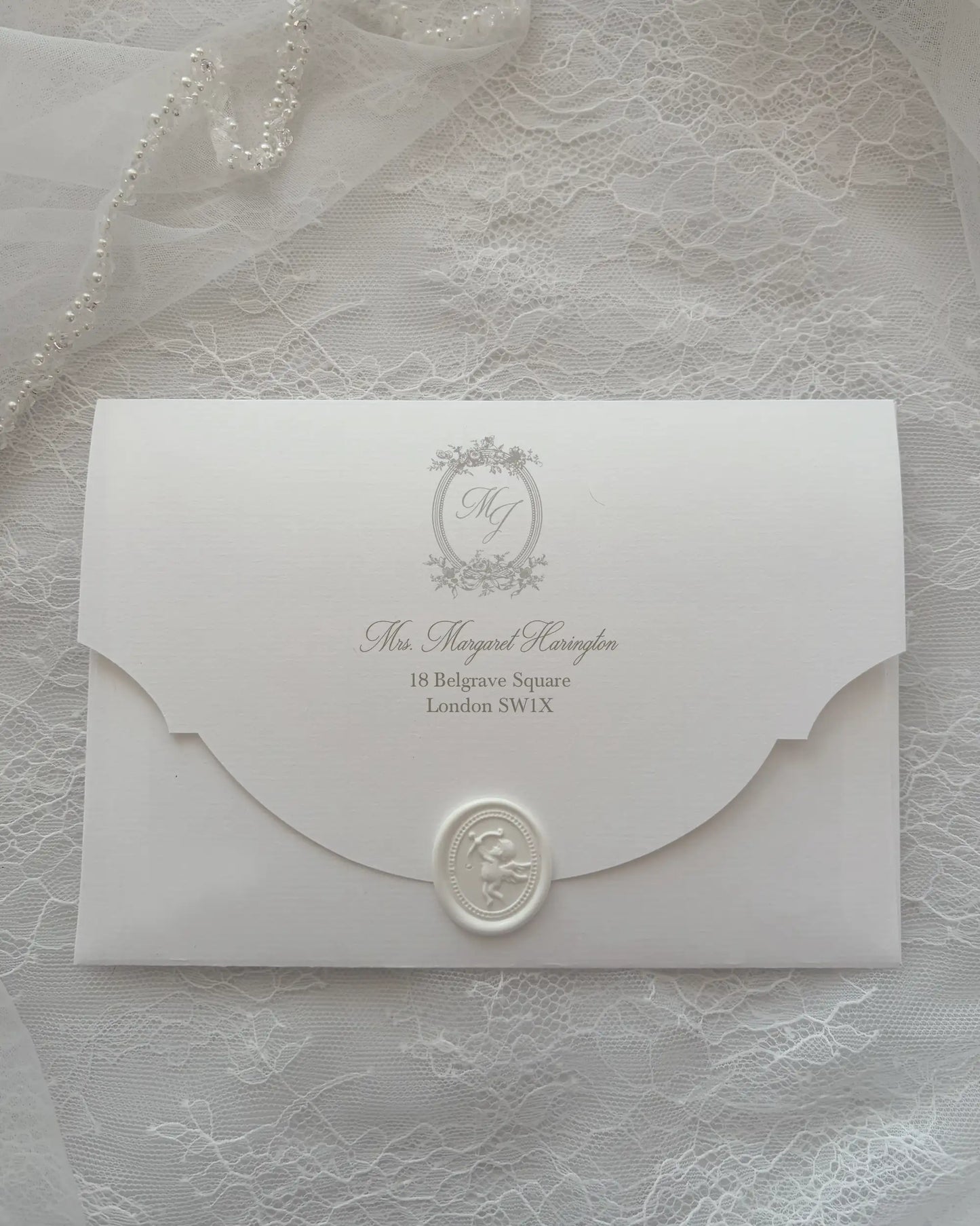 Alyson Lace White envelope with scalloped flap, embossed monogram crest and white wax seal, laid on delicate lace fabric — Polina Perri. 