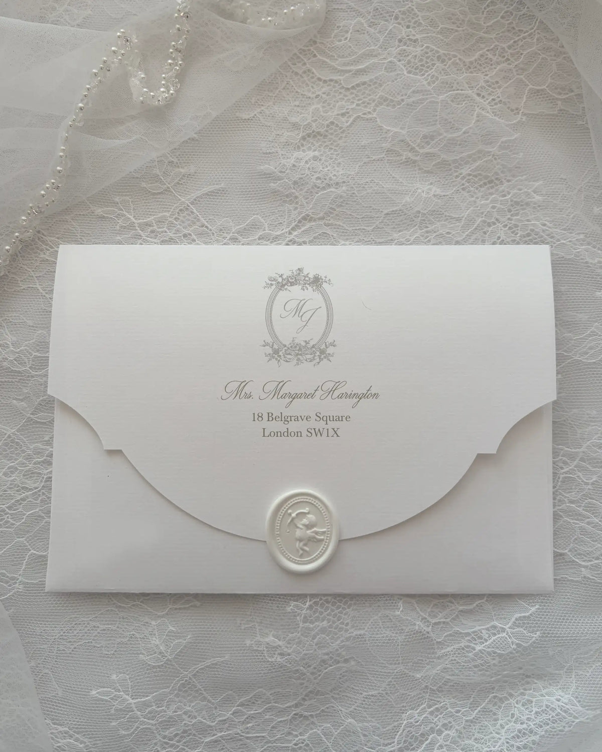 Alyson Lace White envelope with scalloped flap, embossed monogram crest and white wax seal, laid on delicate lace fabric — Polina Perri. 