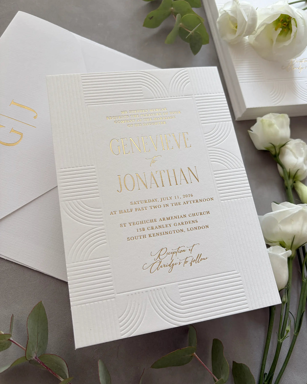Art Deco wedding invitation with embossed wave-pattern and gold foil details arranged in flat lay with white roses and green eucalyptus leaves, matching envelope partially visible — Polina Perri
