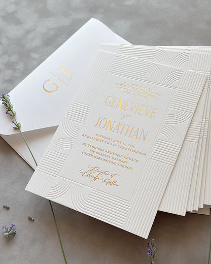 Stack of Art Deco wedding invitations with embossed wave patterns and gold foil lettering, matching envelope and dried lavender sprigs on neutral gray surface — Polina Perri