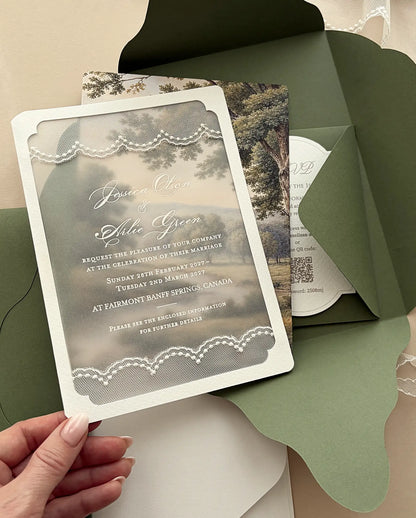 Hand lifting the lace-trimmed vellum wedding invitation out of the olive green four-petal folder, with the landscape print behind
